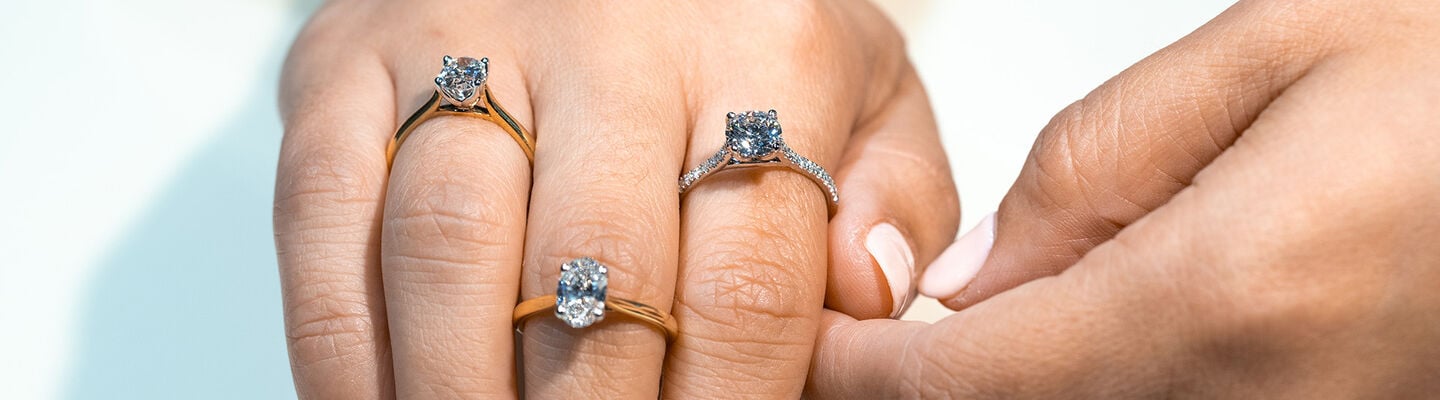 Hand wearing two round-cut and one oval-cut diamond engagement rings in yellow gold and white gold settings.