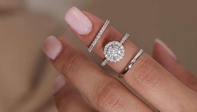 Round halo diamond engagement ring paired with pavé bands and a plain wedding ring on manicured hand.
